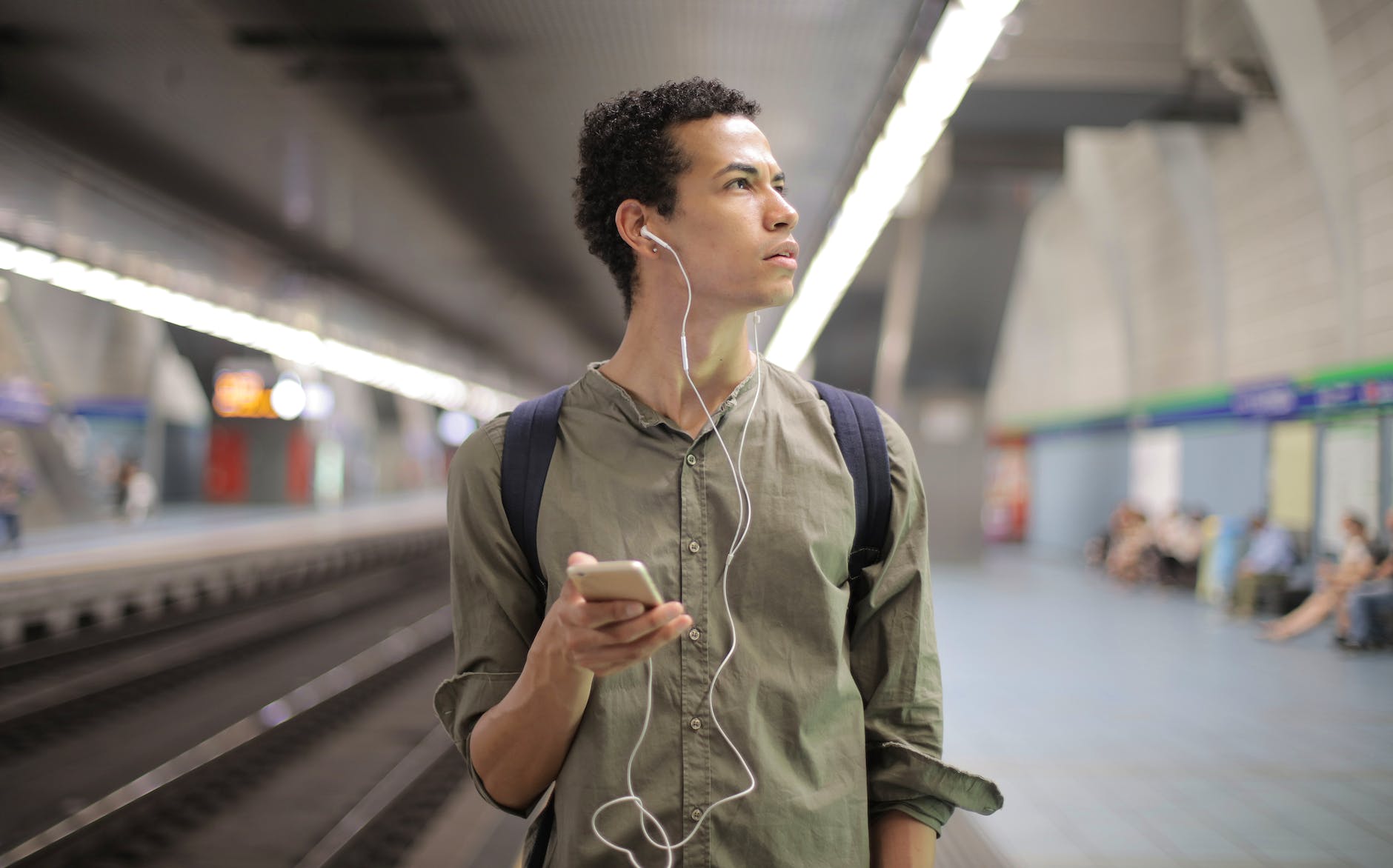 young ethnic man in earbuds listening to music while waiting for transport at contemporary subway station